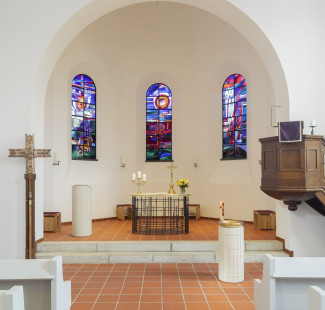 Chorraum der Dreifaltigkeitskirche mit drei bunten Kirchenfenstern im Hintergrund, davor der Altar aus Eisenstreben und der Ambo aus weißem Holz. Rechts von die Kanzel aus dunklem Holz.
