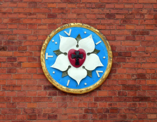 Die Lutherrose besteht aus einem schwarzen Kreuz in der Mitte eines roten Herzens, raum weiße Blütenblätter auf hellblauem Grund, umschlossen wird alles von einem goldenen Ring. 