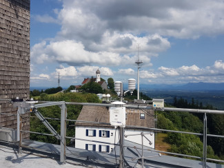 Blick von der Wetterstation auf dem Hohen Peißenberg hinüber zur Wallfahrtskirche. Zu sehen sind technische Messgeräte und blauer Himmel mit Wolken. Das Bild soll zum Gemeindeausflug auf die Wetterstation einladen. 